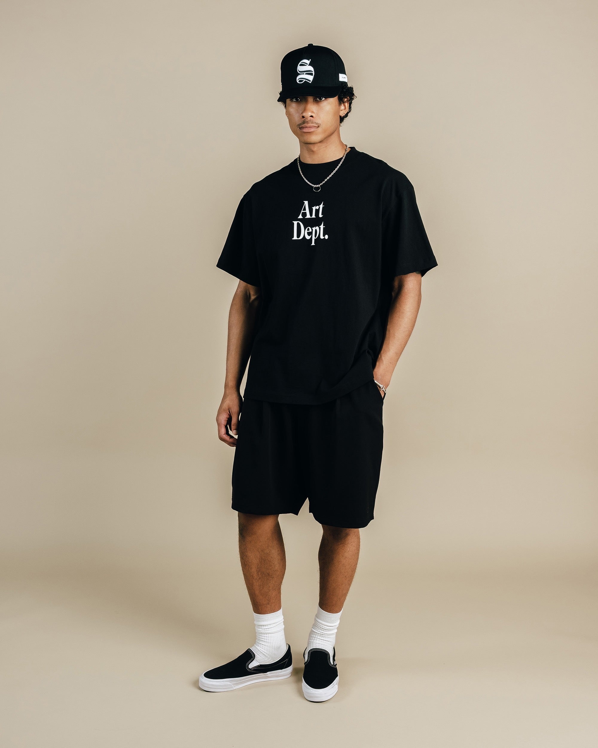 Person wearing a black 'Art Dept.' t-shirt, black shorts, and a black cap on a beige background