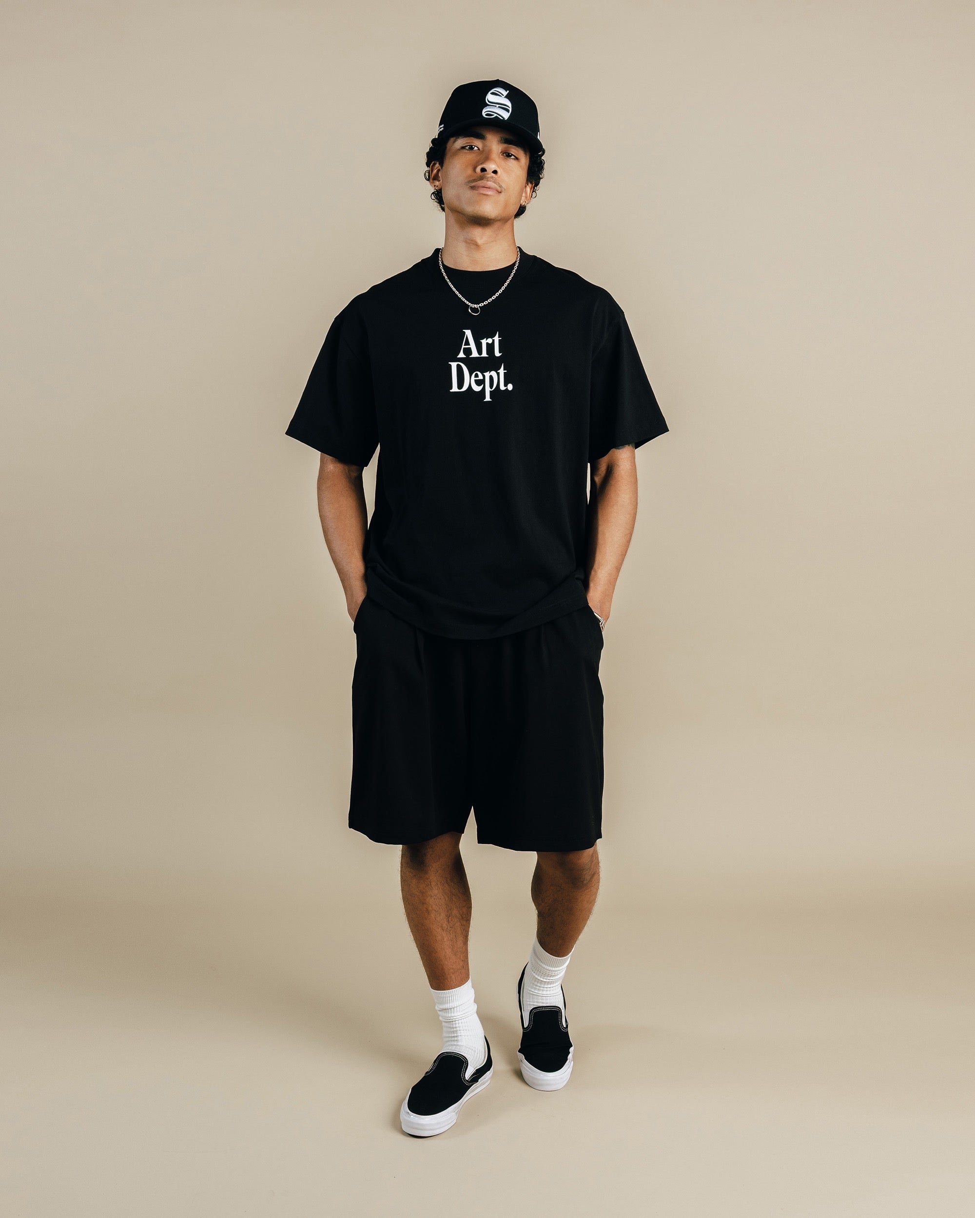 Person wearing a black 'Art Dept.' t-shirt, black shorts, and a black cap on a beige background