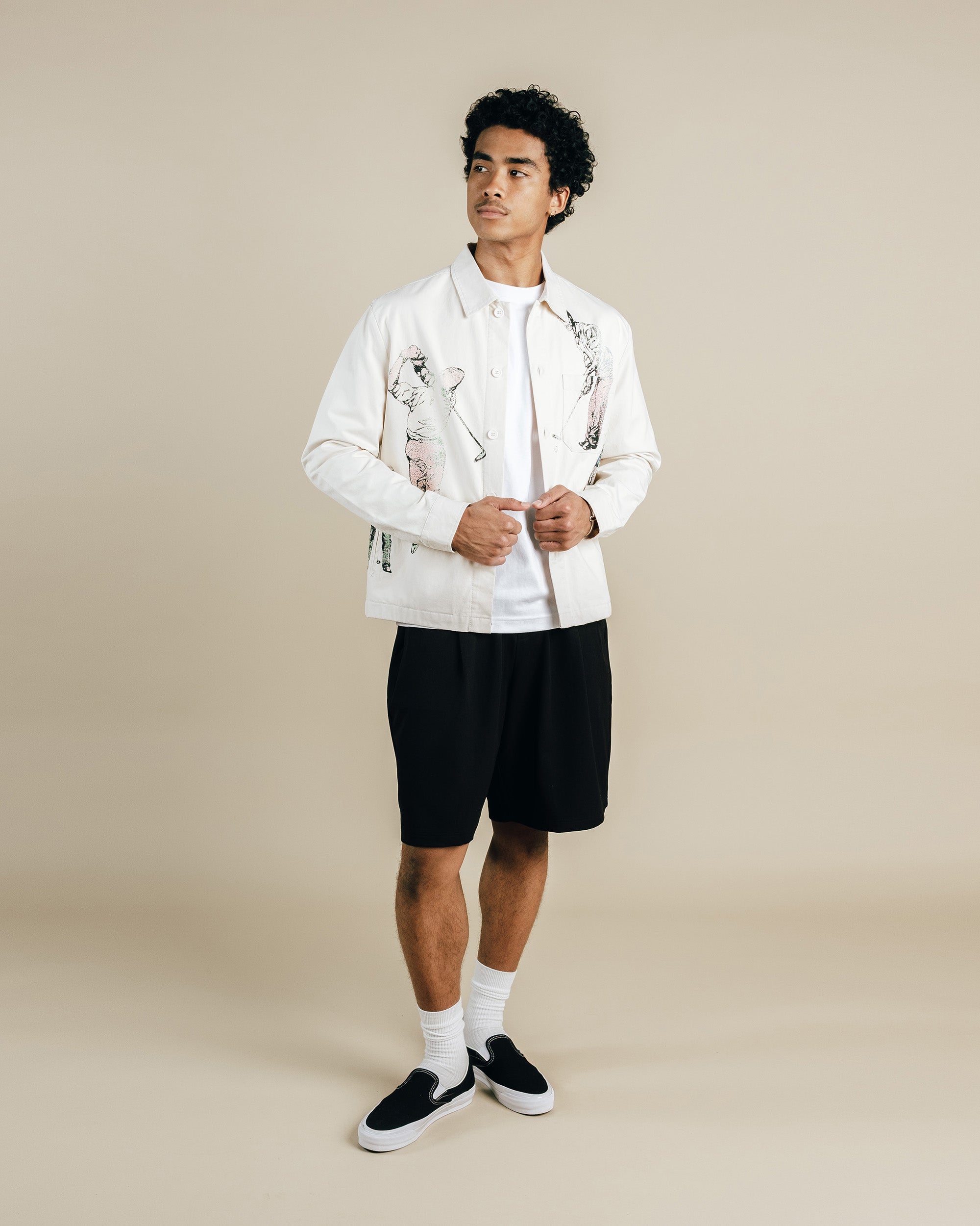 Man wearing a white jacket with black patterns, white shirt, black shorts, and black sneakers on a beige background