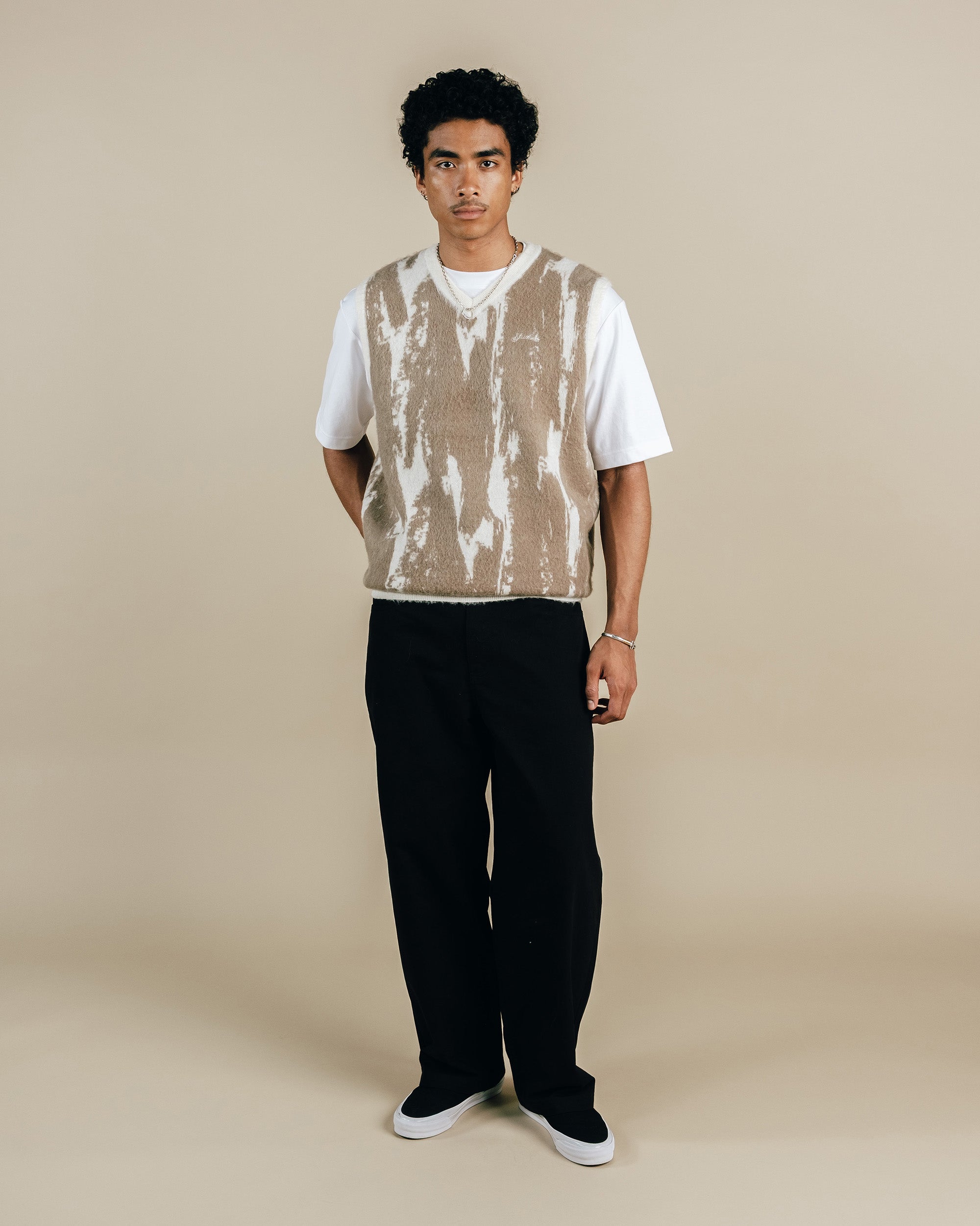 Person wearing a patterned vest over a white shirt with black pants on a beige background