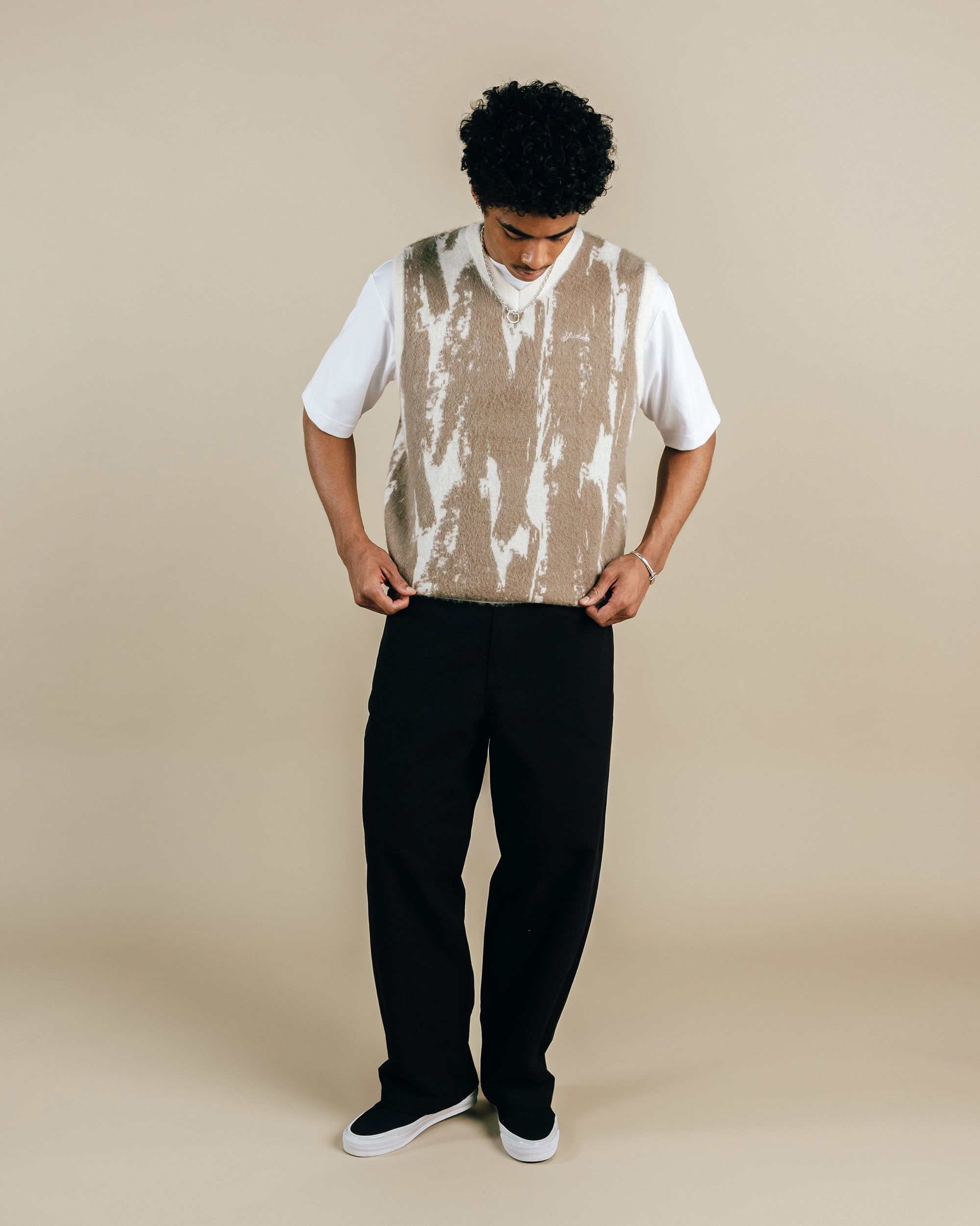 Person wearing a white t-shirt with a beige tie-dye vest and black pants on a beige background