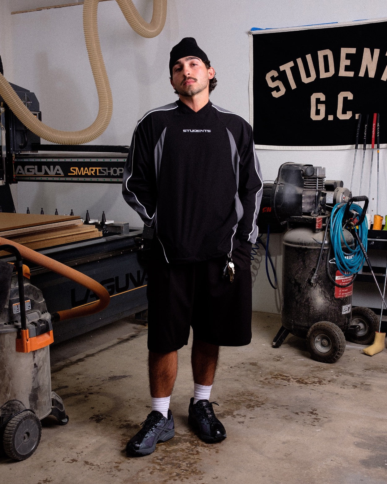 Man standing in a workshop wearing a black and gray sweatshirt with 'STUDENTS' branding.