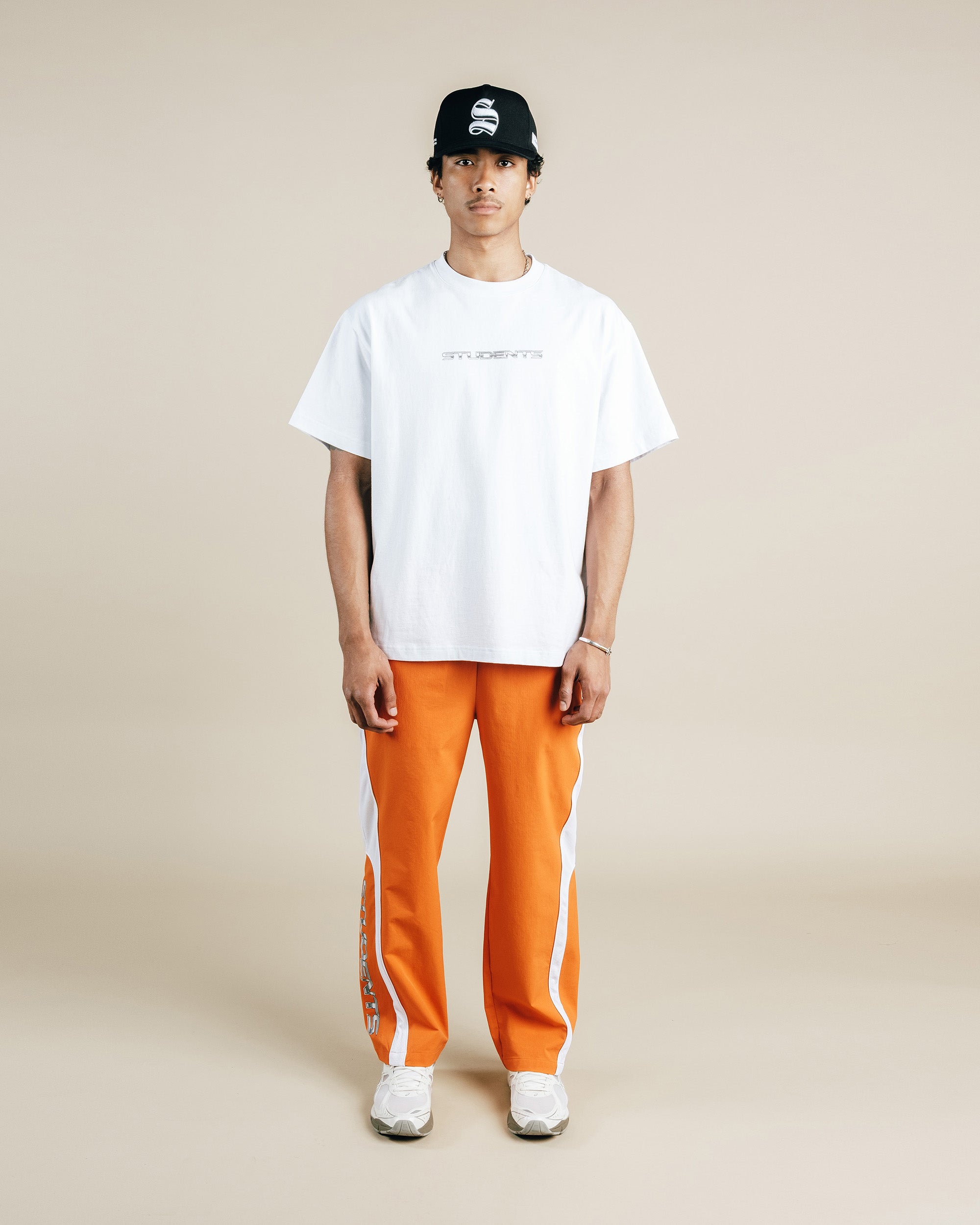 Person wearing a white t-shirt, orange pants, and a black cap on a beige background