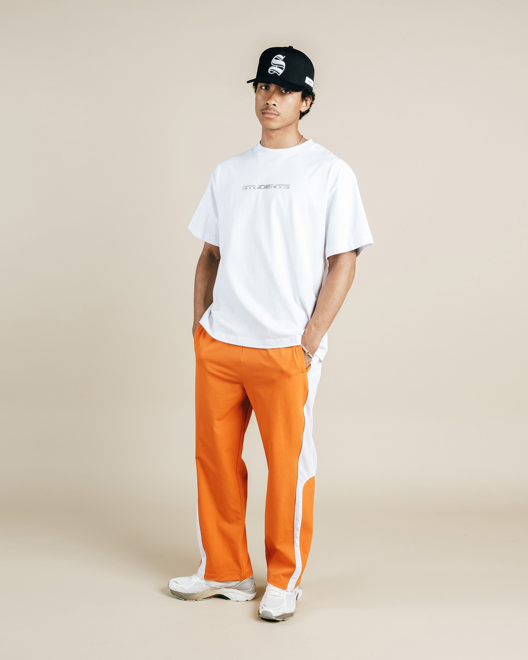 Person wearing a white t-shirt, orange pants with a white stripe, and a black cap on a beige background