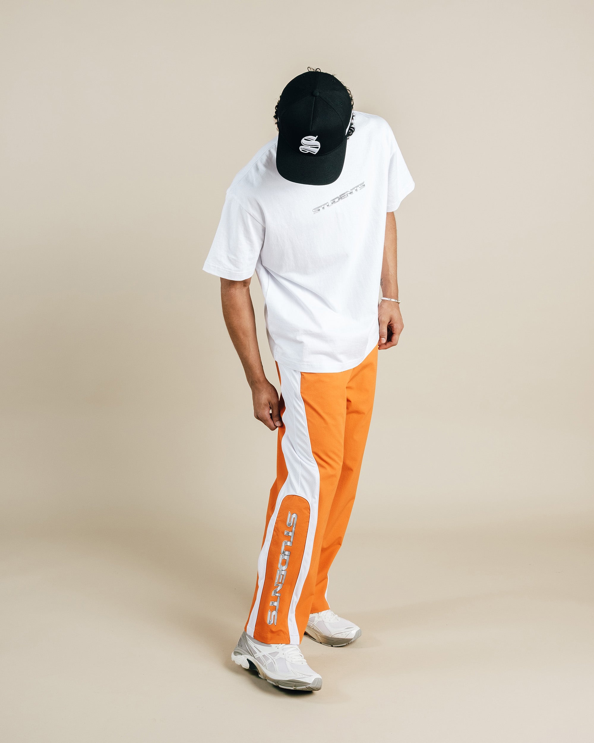 Person wearing a white t-shirt, black cap, and orange pants with a brand logo on a beige background