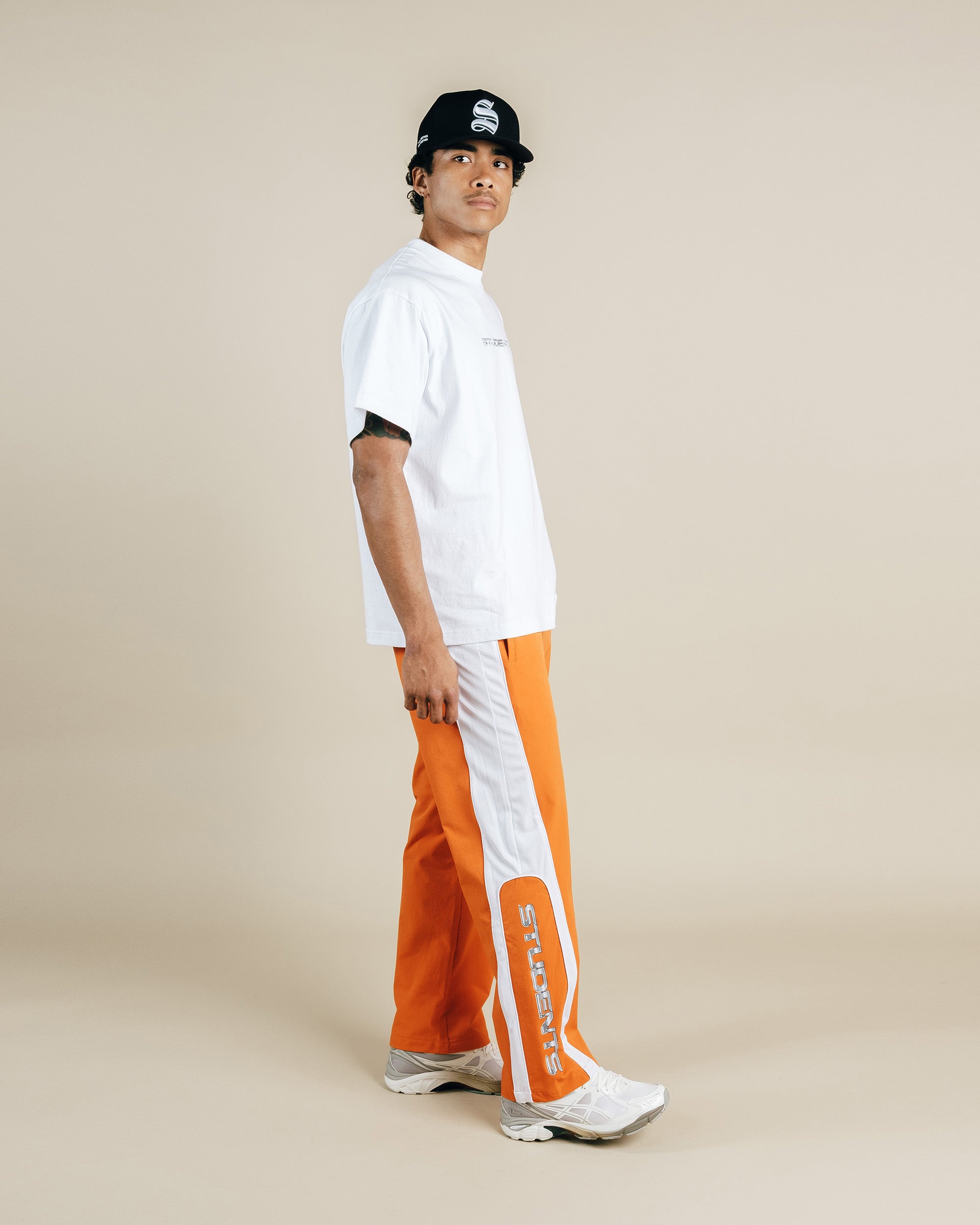 Person wearing a white shirt, orange pants with white stripes, and a black cap on a beige background