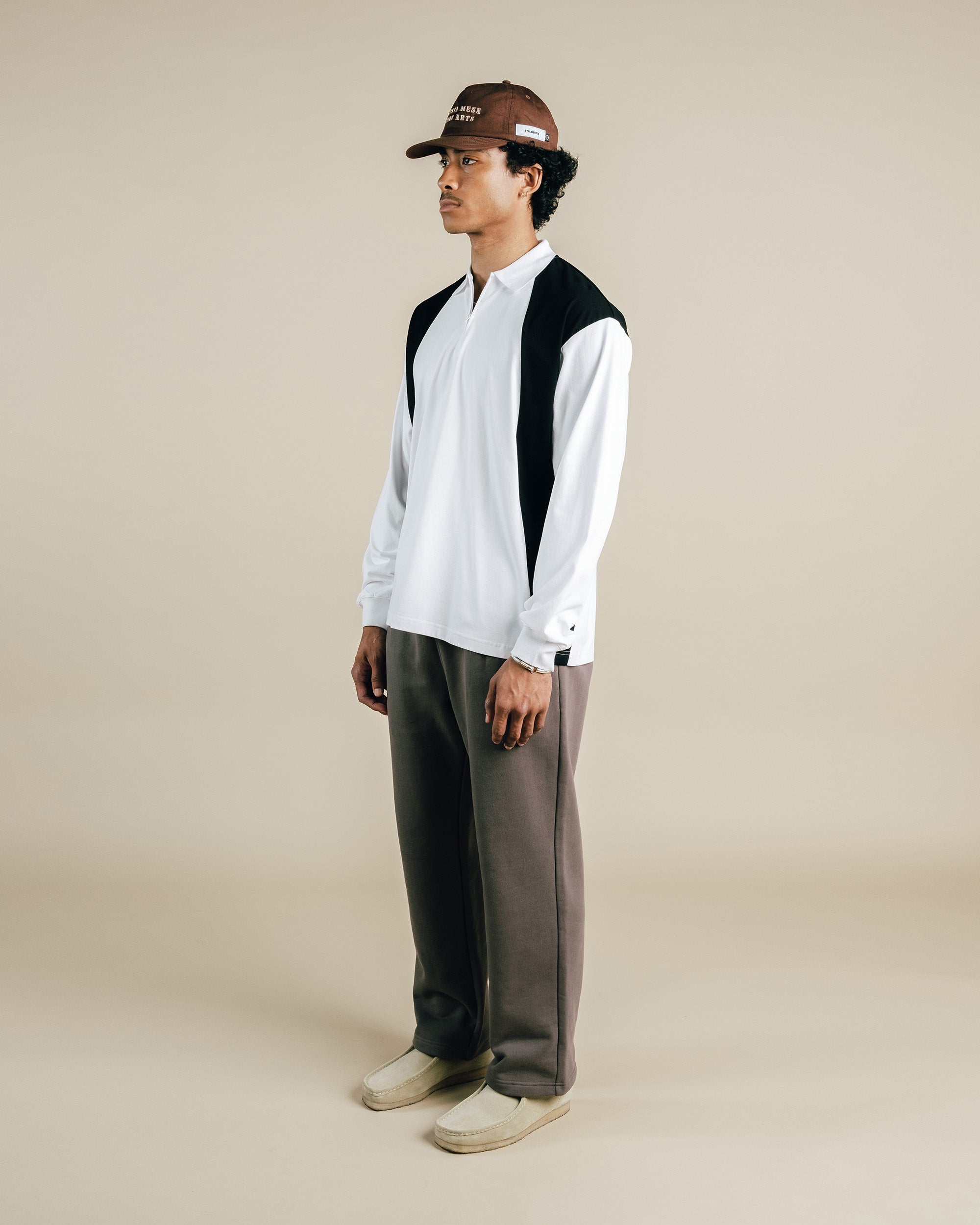 Person wearing a black and white shirt with a vest, brown pants, and a cap on a beige background