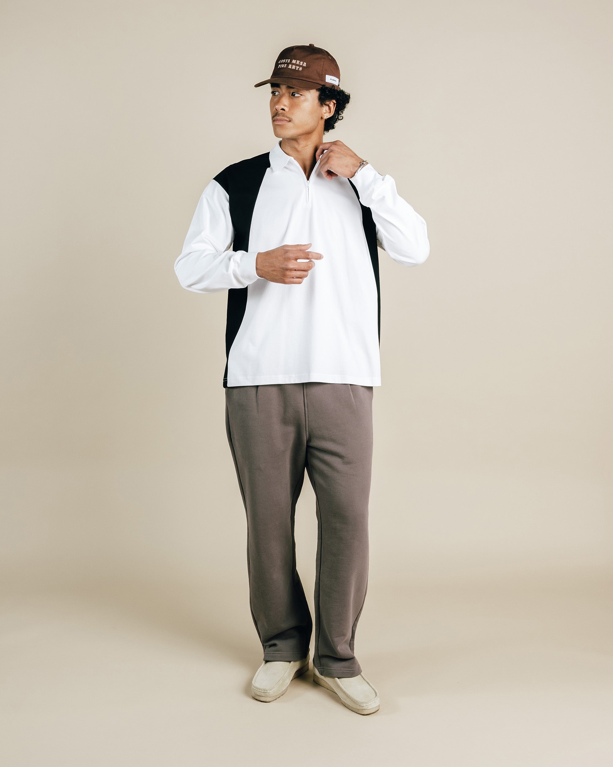 Man wearing a black and white long sleeve shirt, brown pants, and a cap on a beige background