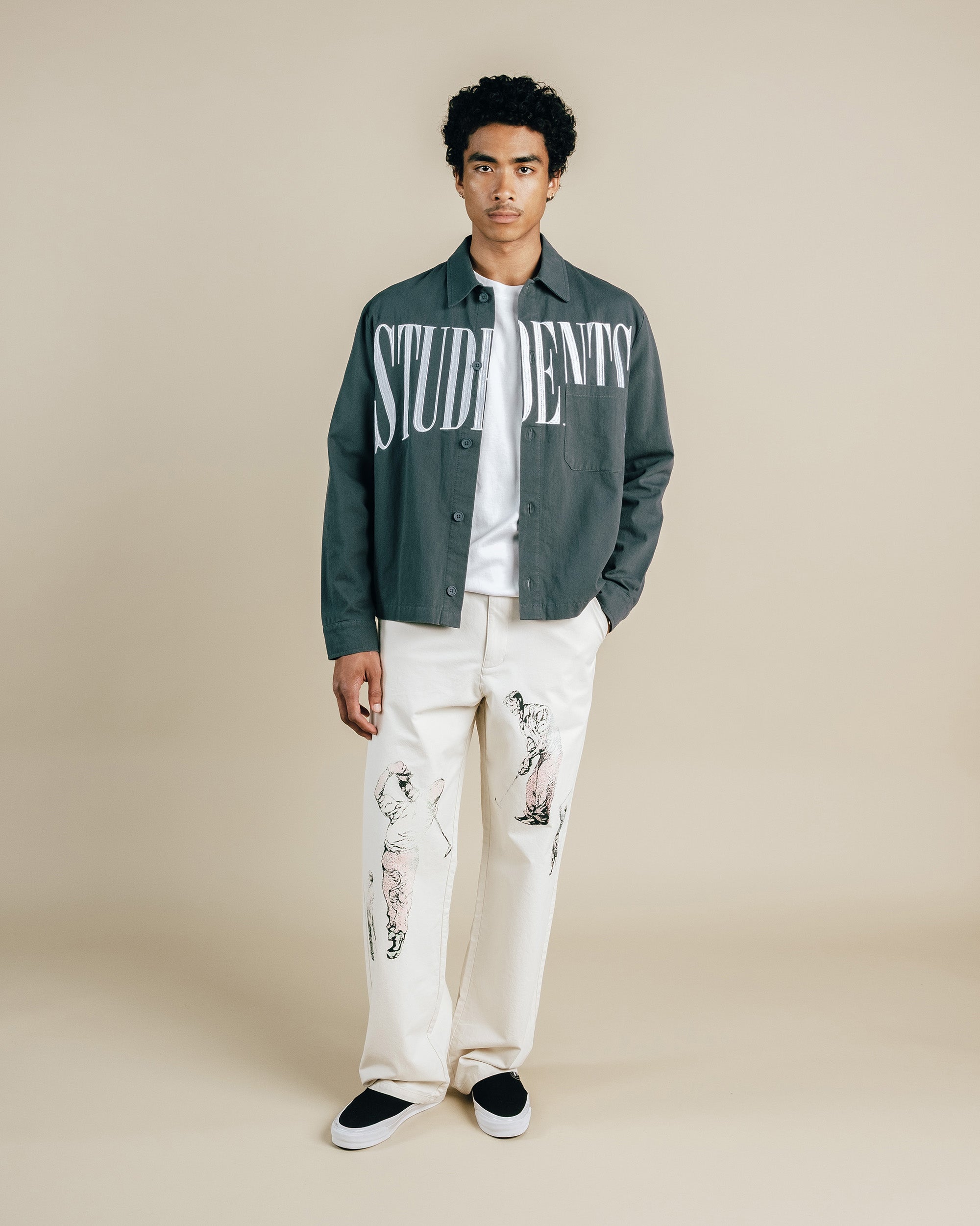 Man wearing a green jacket with 'STUDENTS' text, white shirt, and white pants with black patterns on a beige background