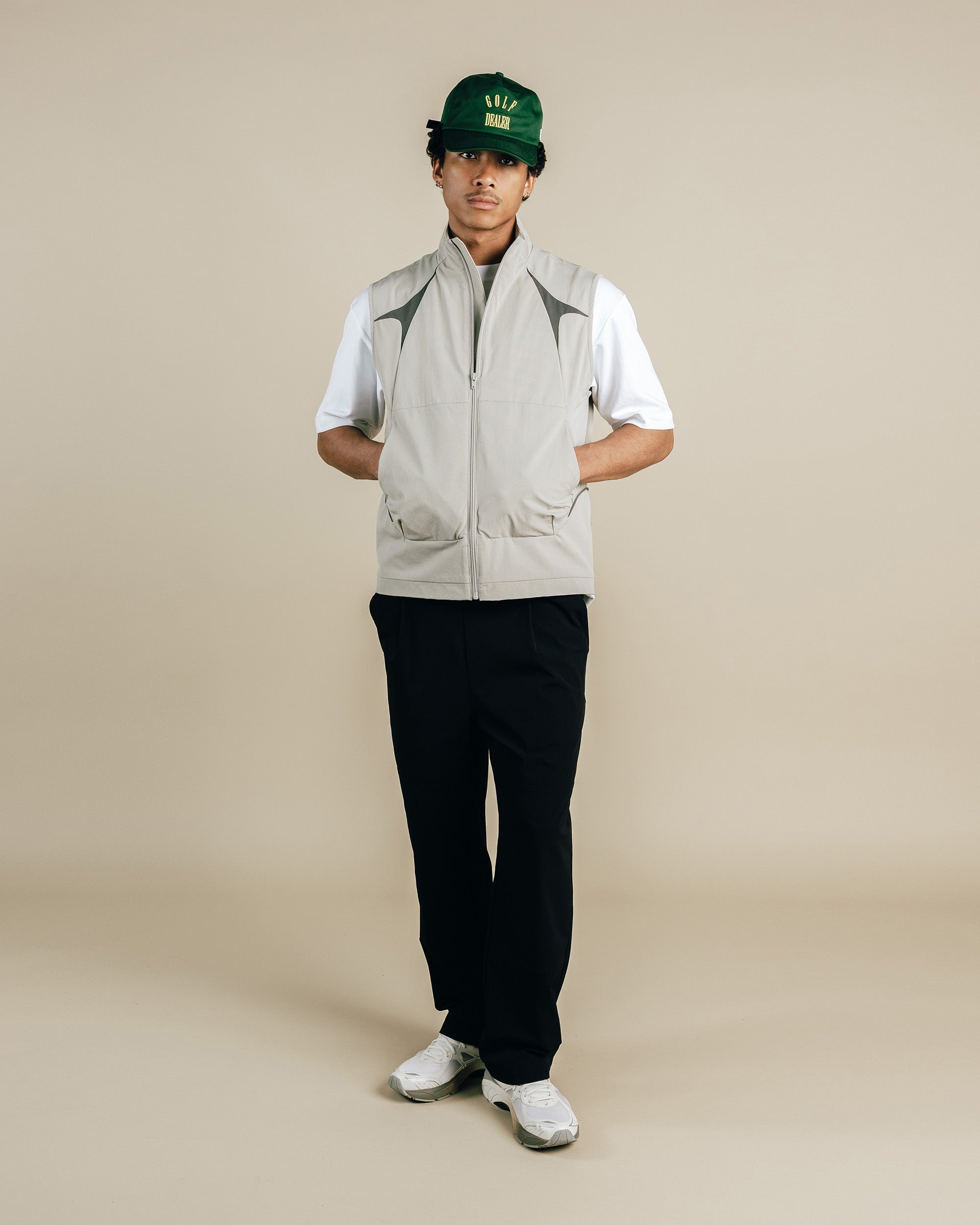 Person wearing a light gray vest, black pants, white shirt, and green cap on a beige background