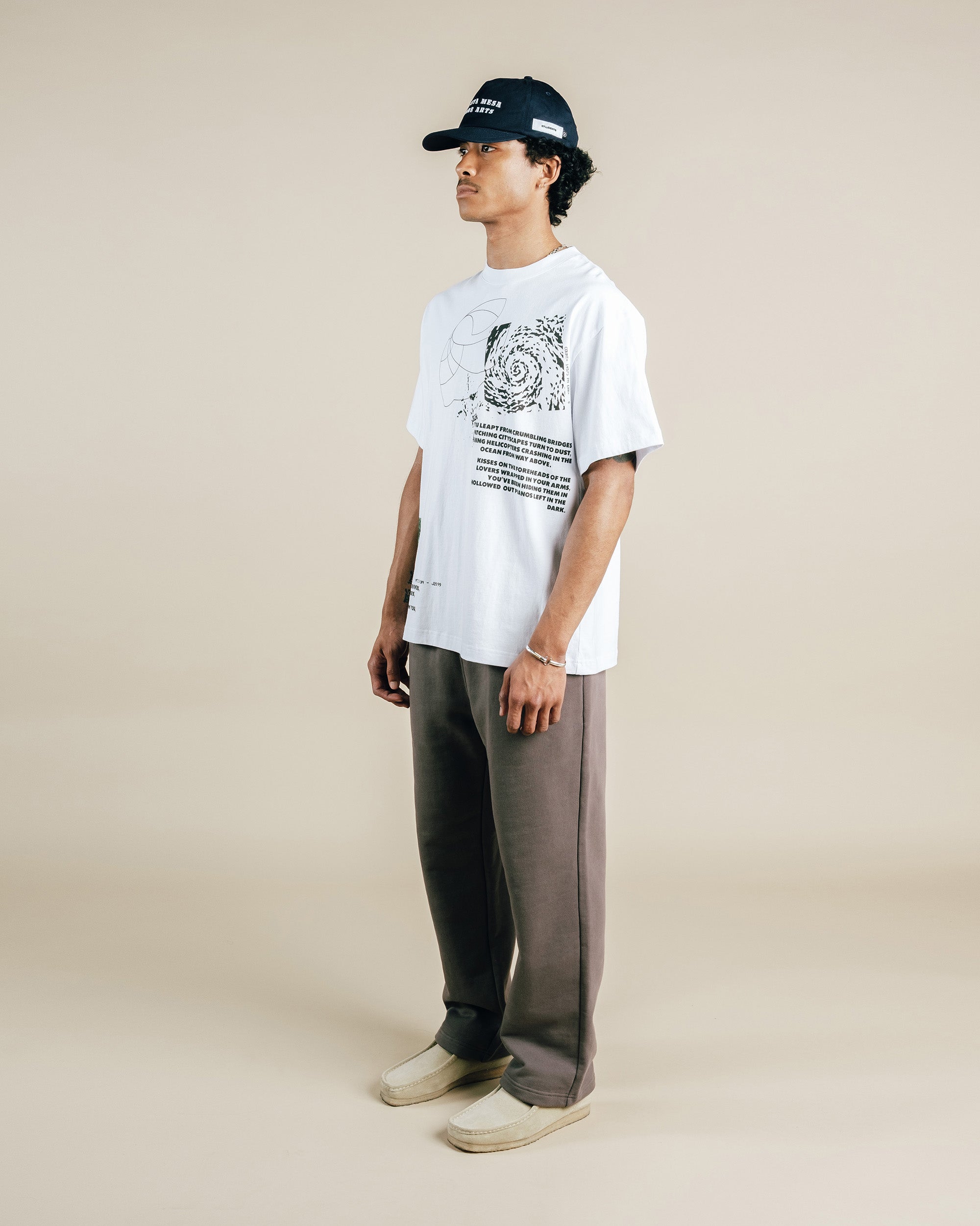 Person wearing a white t-shirt with graphic design, dark pants, and a cap on a beige background