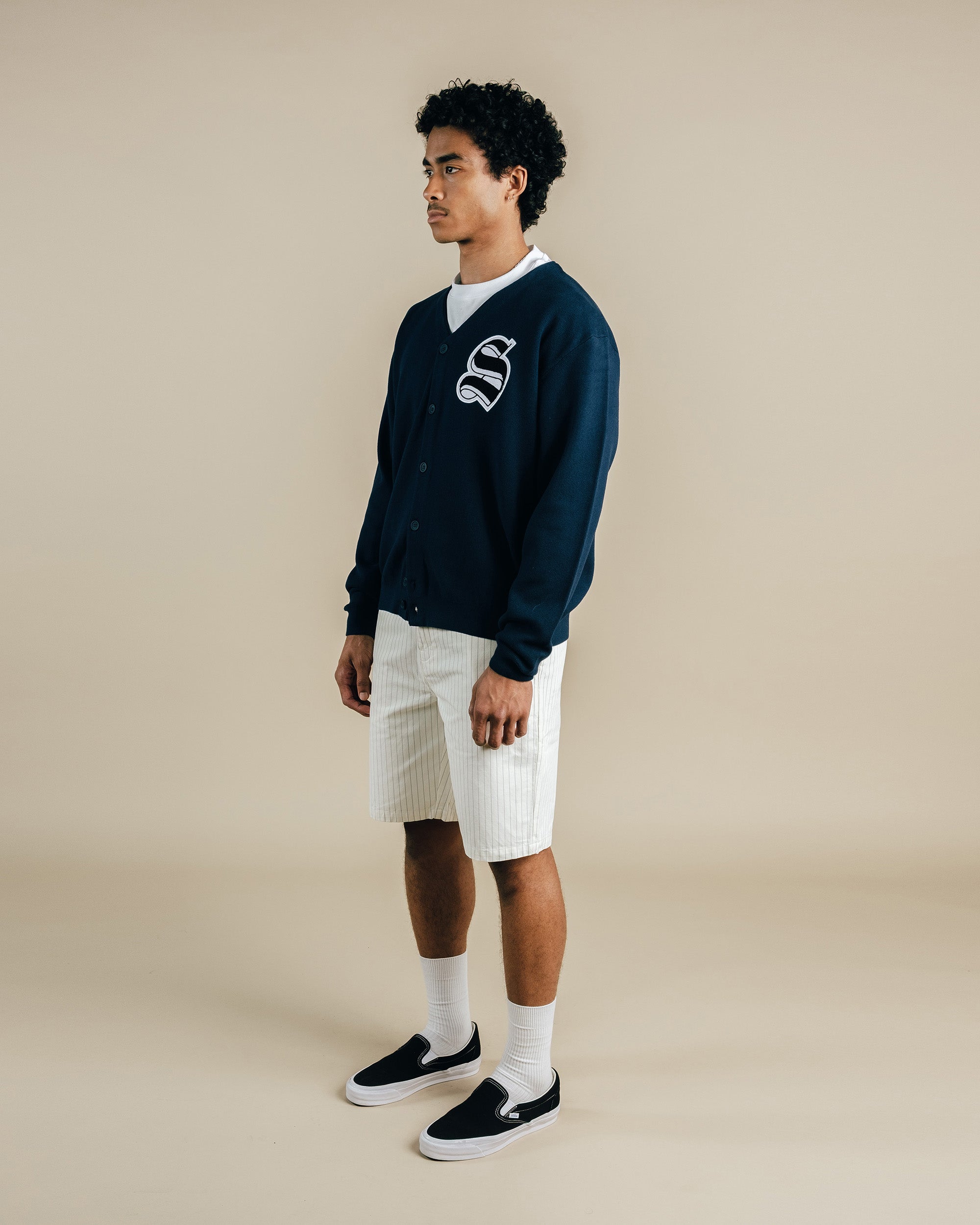 Man wearing a navy blue varsity jacket with a logo, white shorts, and black sneakers on a beige background