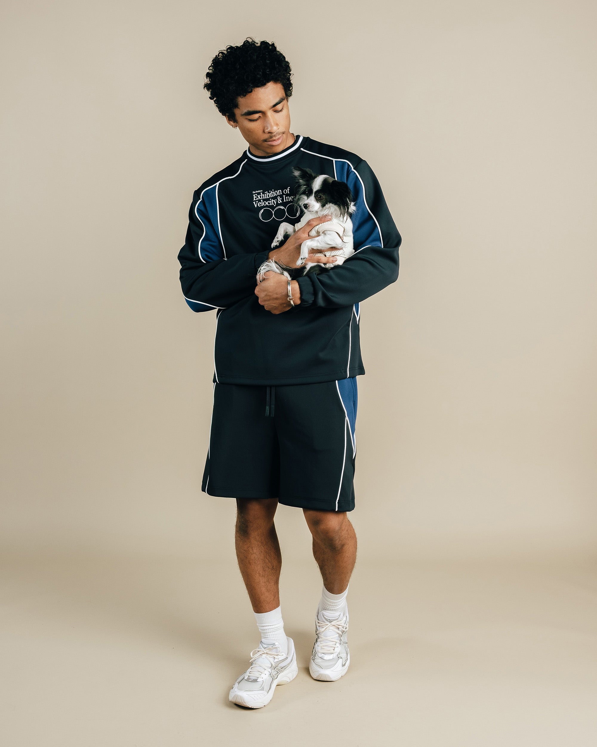 Man holding a small dog wearing a matching black and blue outfit against a beige background