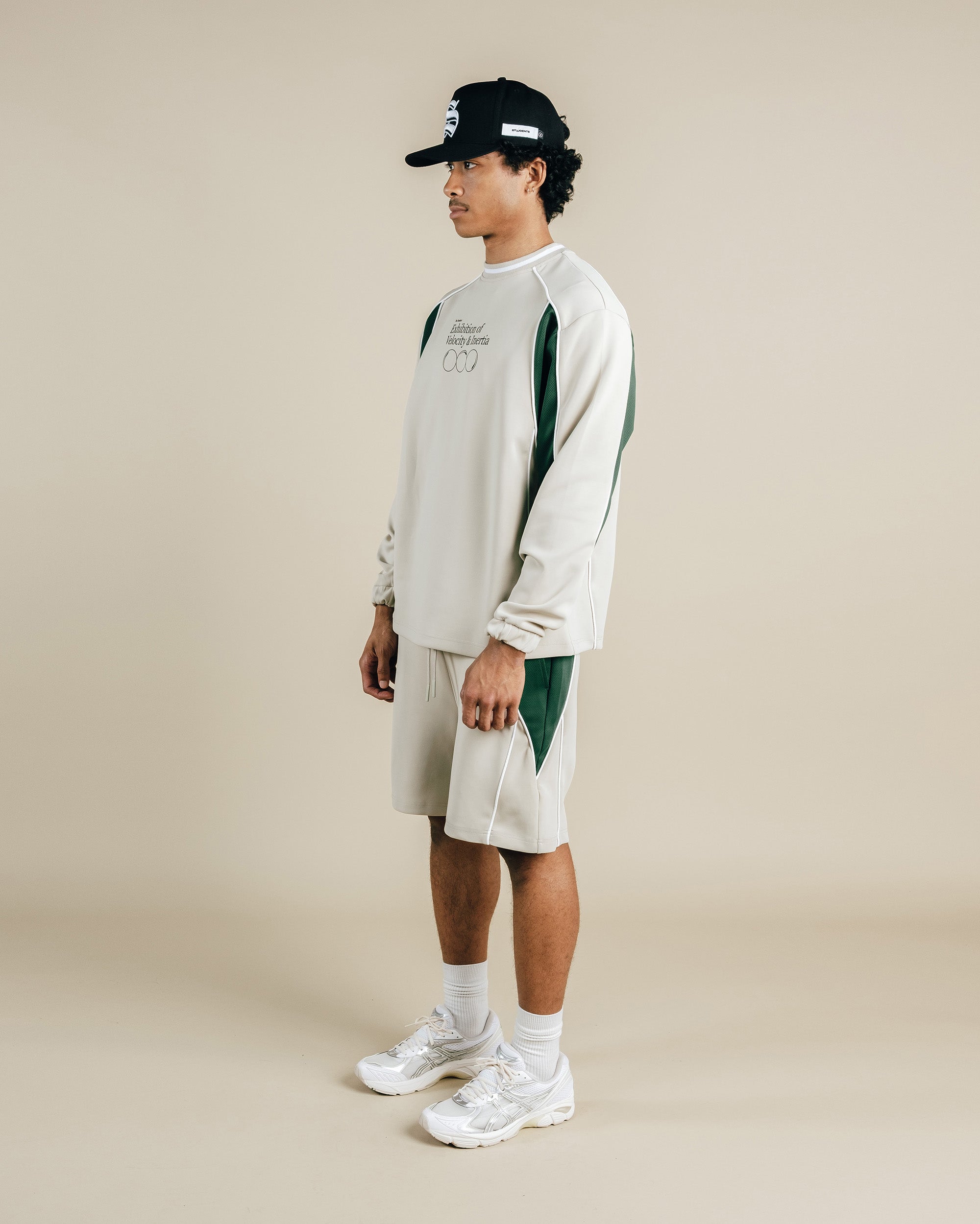 Person wearing a light gray sweatshirt with green accents, beige shorts, and white sneakers on a beige background
