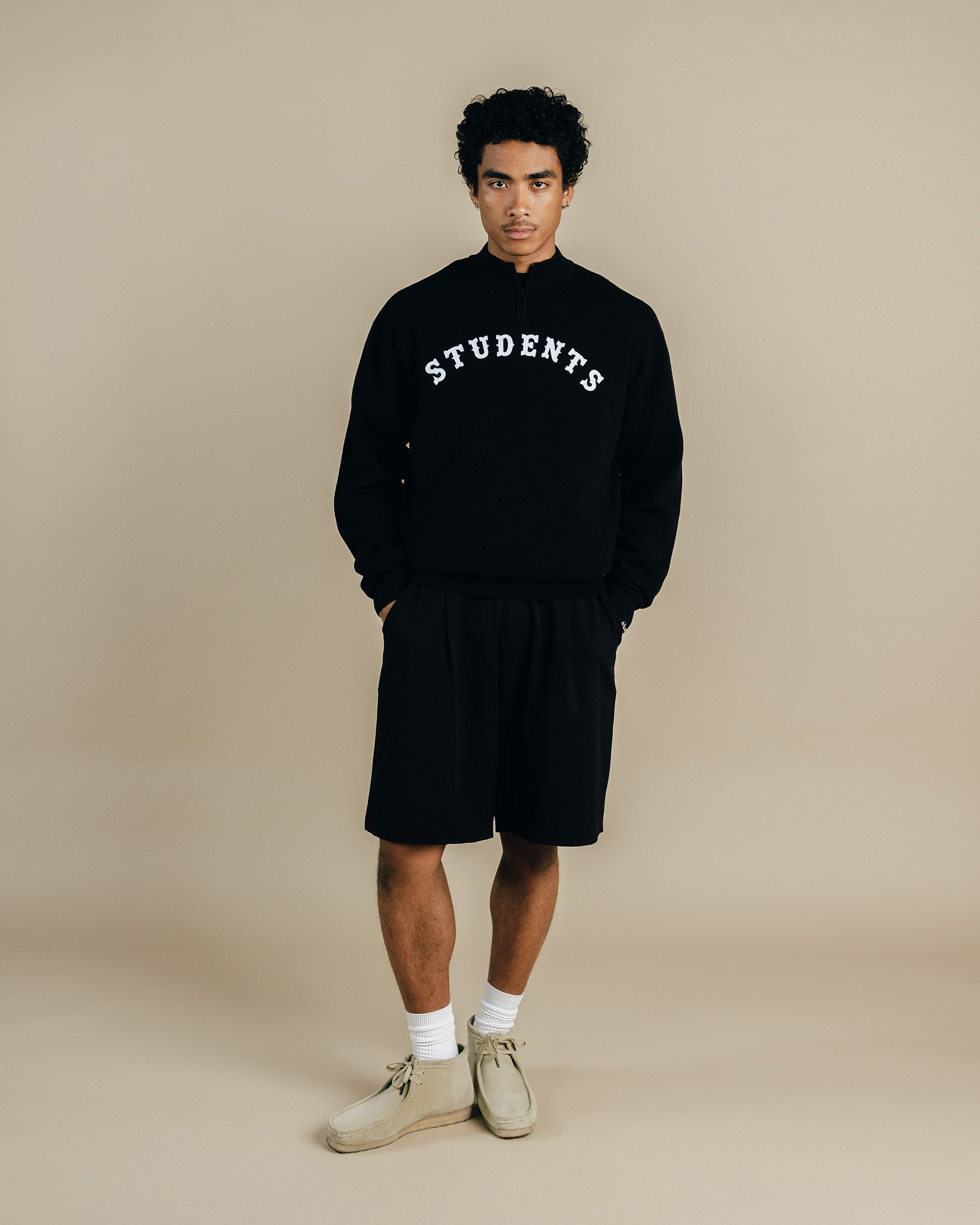 Person wearing a black 'STUDENTS' sweatshirt and shorts on a beige background
