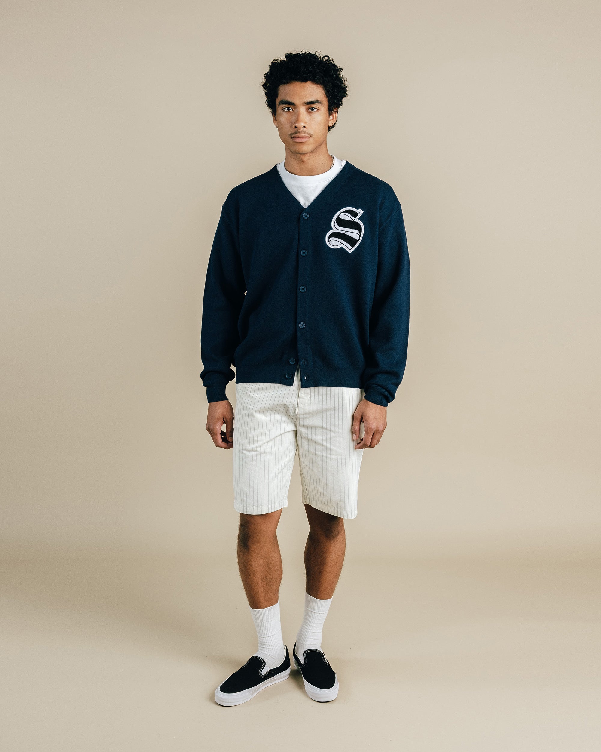 Person wearing a navy cardigan with a logo, white shirt, shorts, and black shoes on a beige background