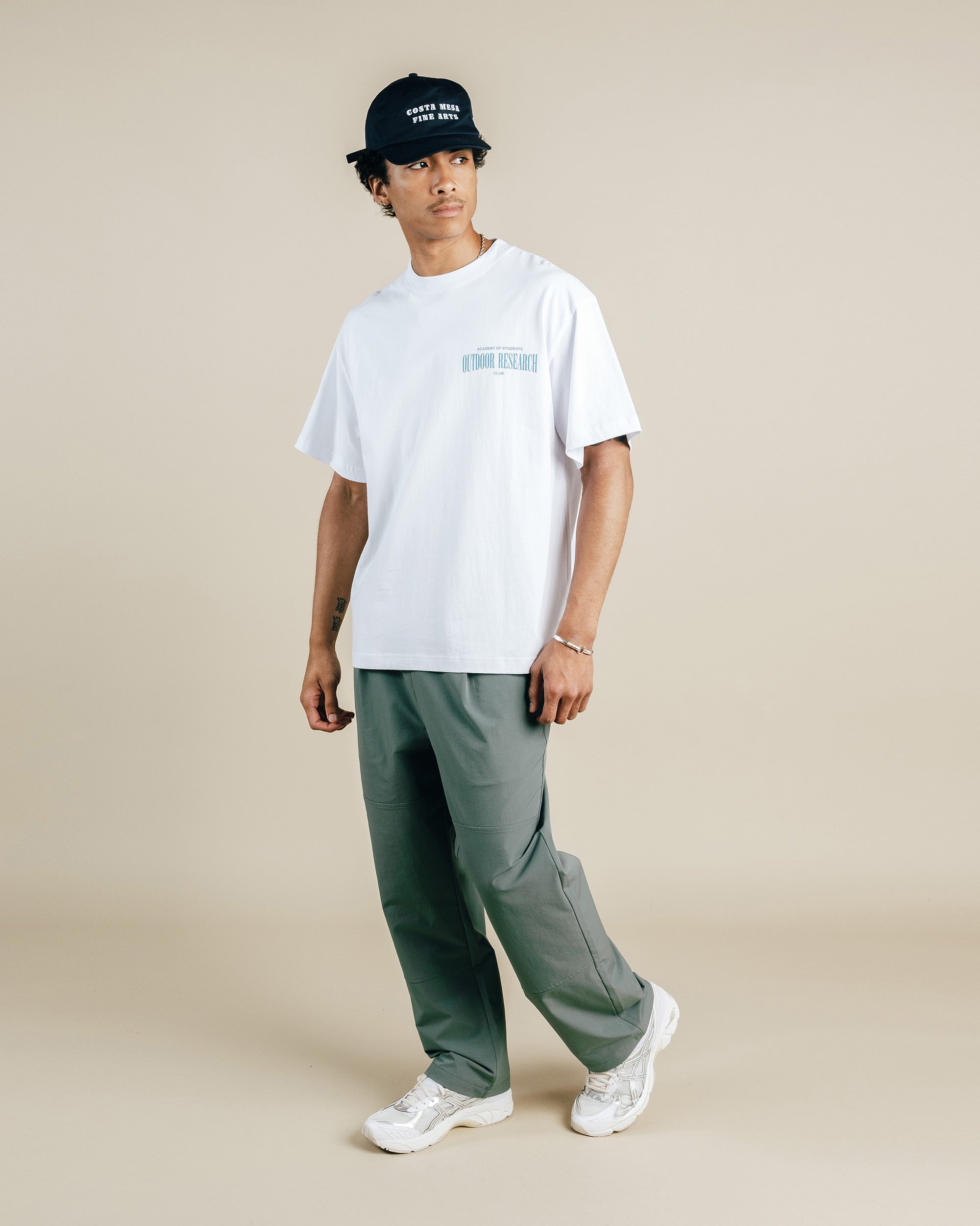 Person wearing a white t-shirt, green pants, and a black cap on a beige background