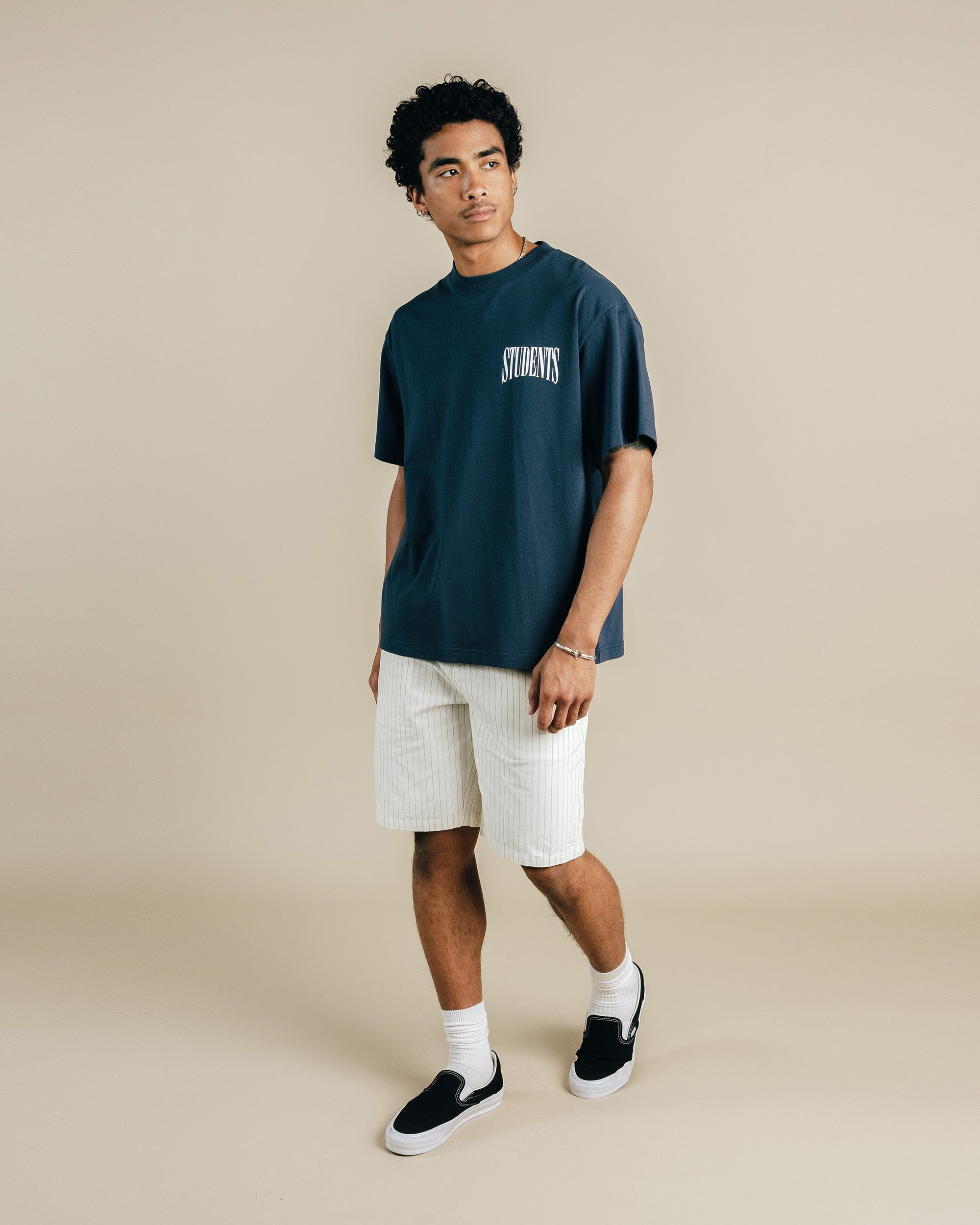 Person wearing a navy blue t-shirt with text, white shorts, and black shoes on a beige background