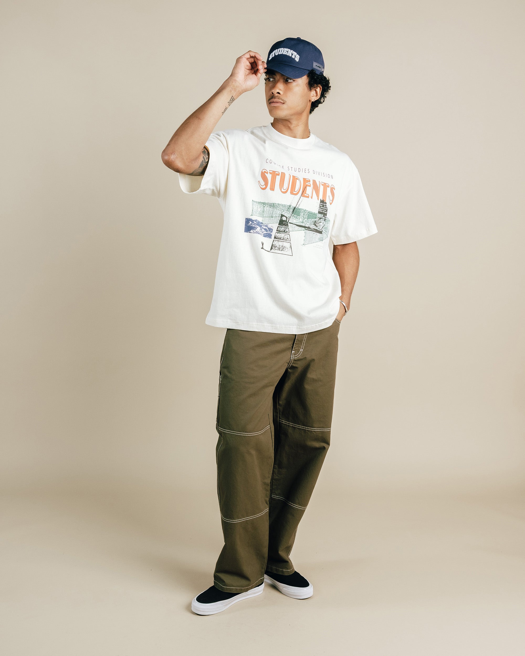 Person wearing a nude t-shirt with graphics and text, green pants, and a navy cap on a beige background