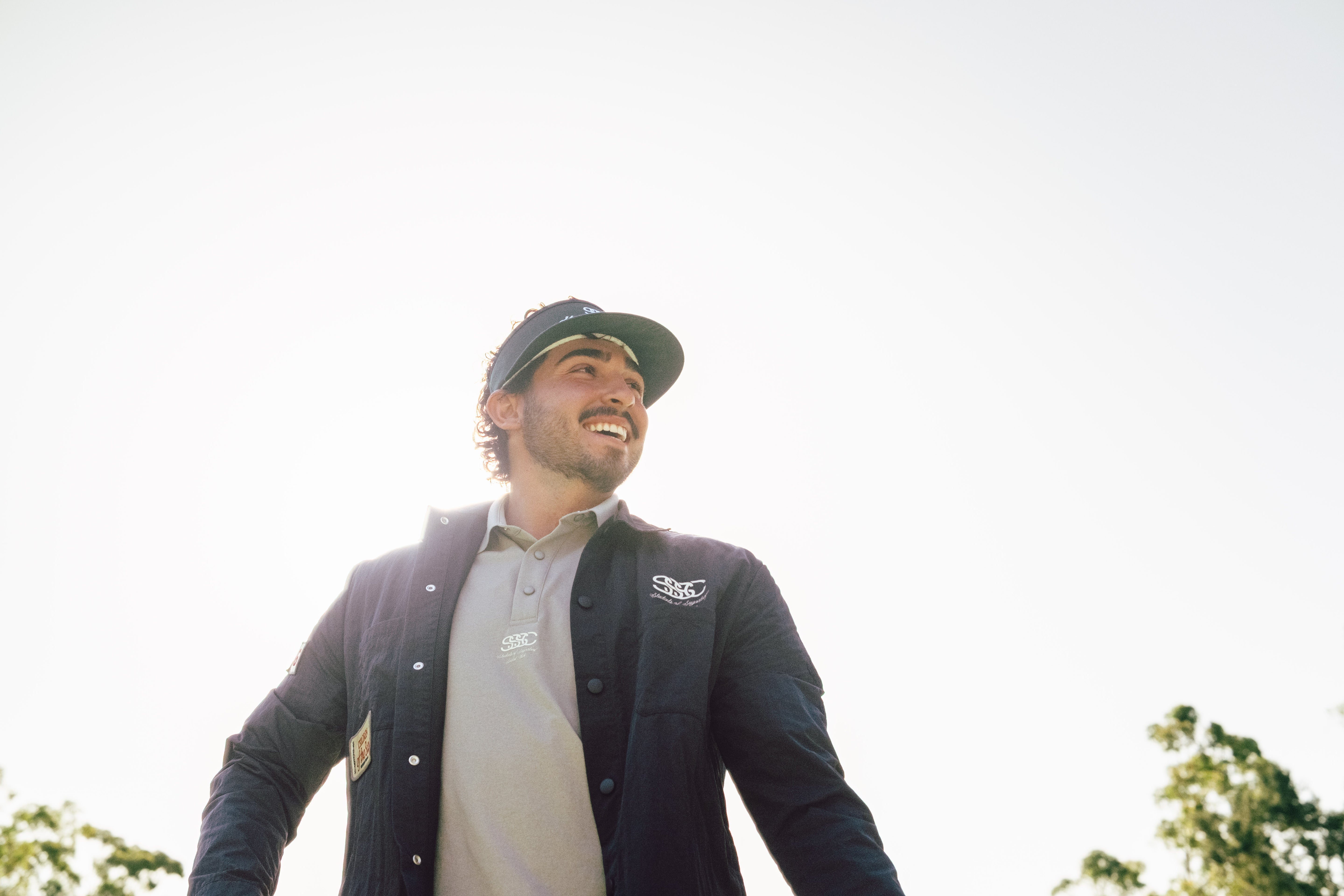 Man wearing a dark jacket with a logo, smiling outdoors.