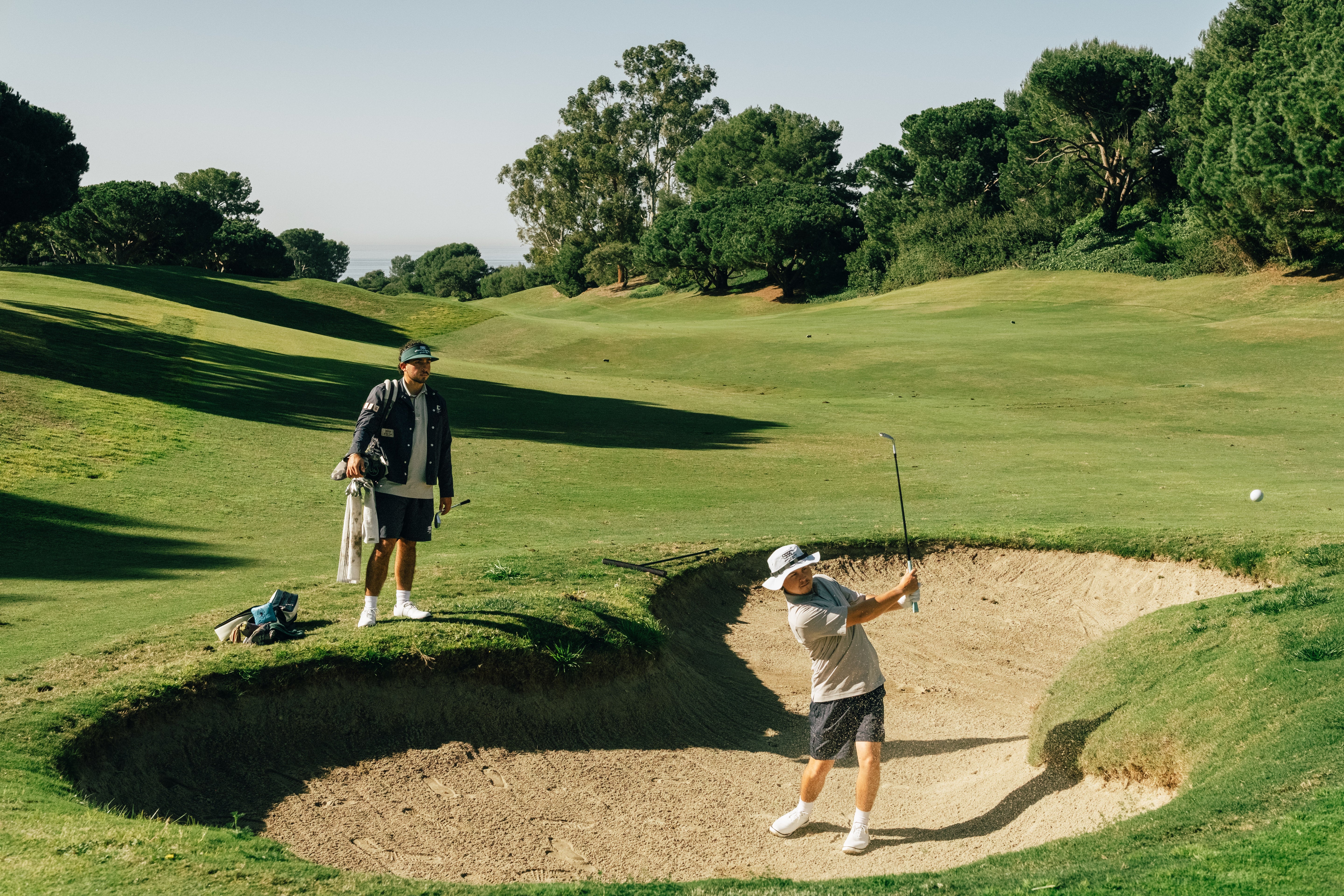Two golfers on a green course with one hitting a ball from a sand trap.