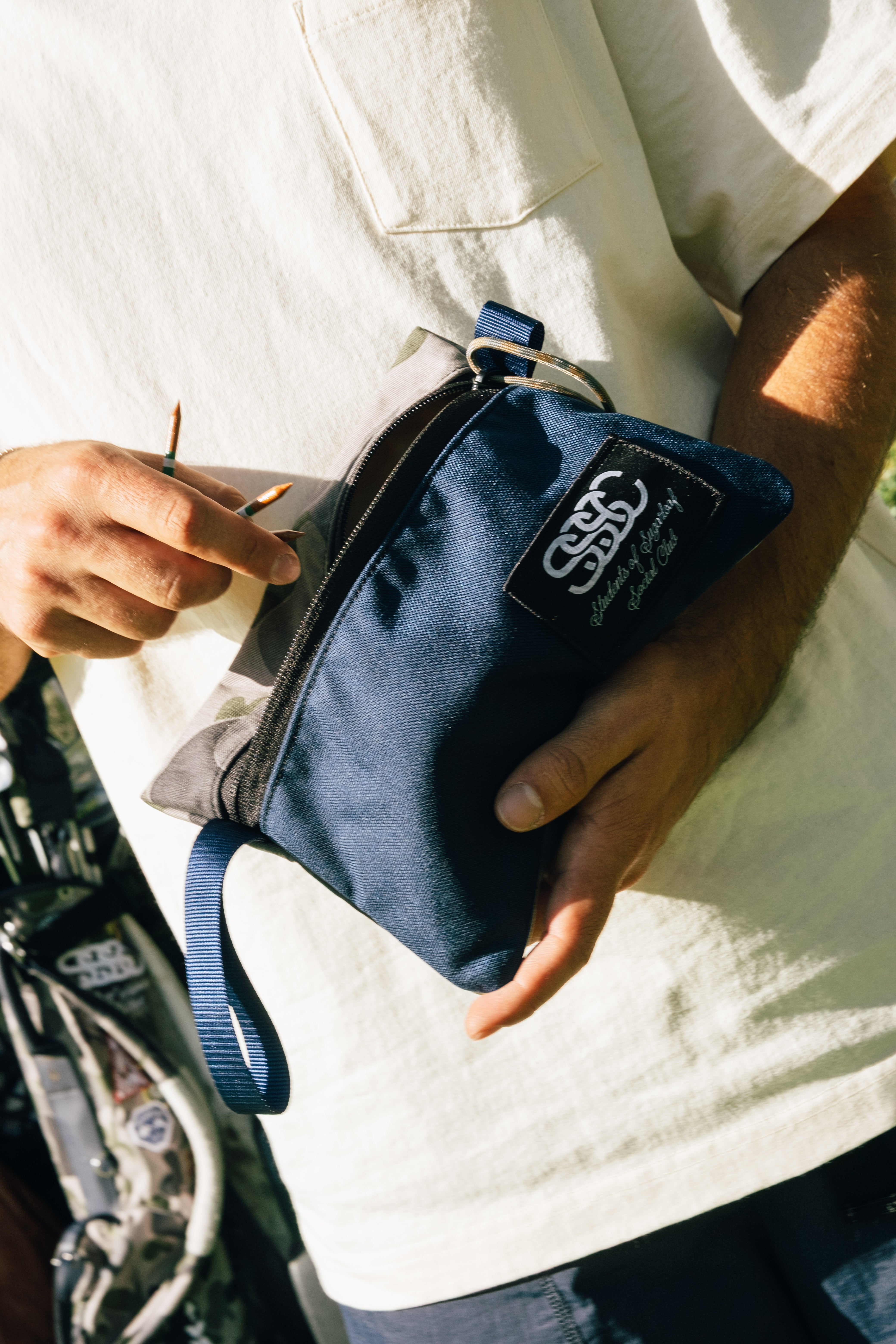 Person holding a navy blue pouch with a brand logo, wearing a white shirt.